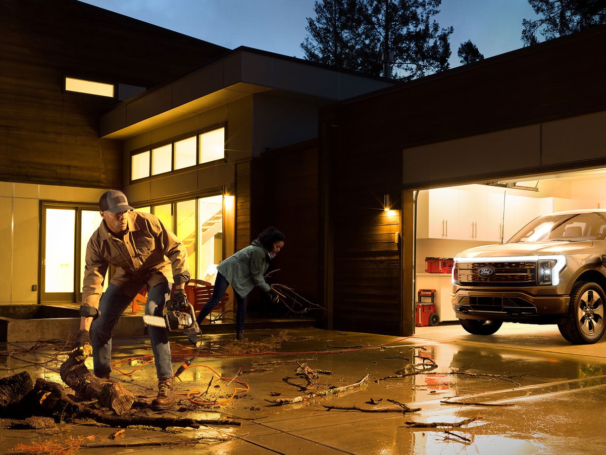 A man and woman cleaning up their driveway from a storm while their F-150 Lightning is charging the house.