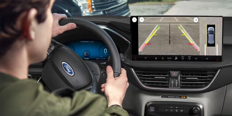 Person in a car looking at a dashboard screen displaying a rear-view camera with parking lines and an overhead parking assist view.