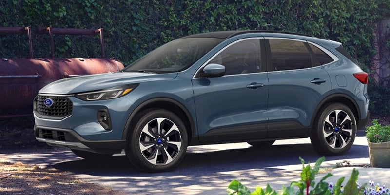 A slate blue Ford Escape SUV parked on pavement with green foliage and a rusty smoker in the background.