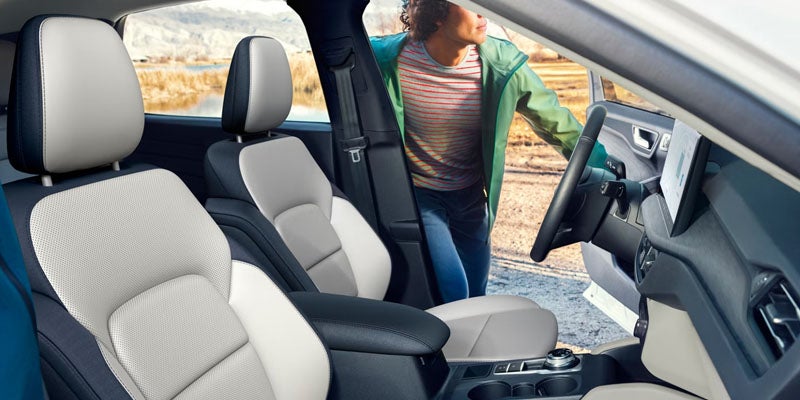 Car interior with two-tone light grey and dark blue seats, and a person in a green jacket leaning into the open driver's door.