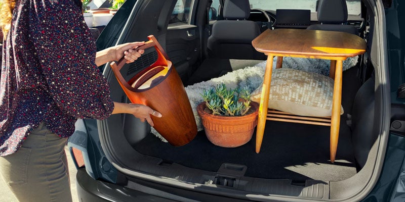 Person loads a brown bucket, potted plant, and small wooden table into the open trunk of an SUV.