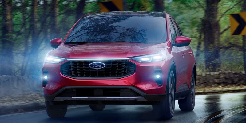 Red Ford SUV driving on a wet road, headlights glowing, with blurred trees and a yellow sign in the background.