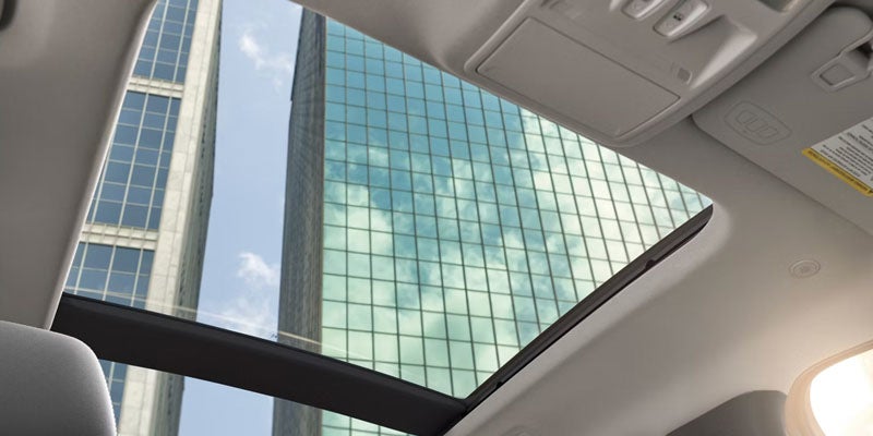View through a vehicle's sunroof at tall city buildings with reflective glass windows, showing cloud reflections.